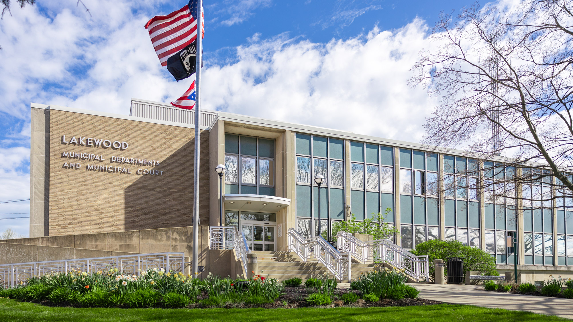 Lakewood City Hall exterior photo Tan brick building with green exterior walls and windows. Blue sky with clouds in the background, Flagpole in the foreground.