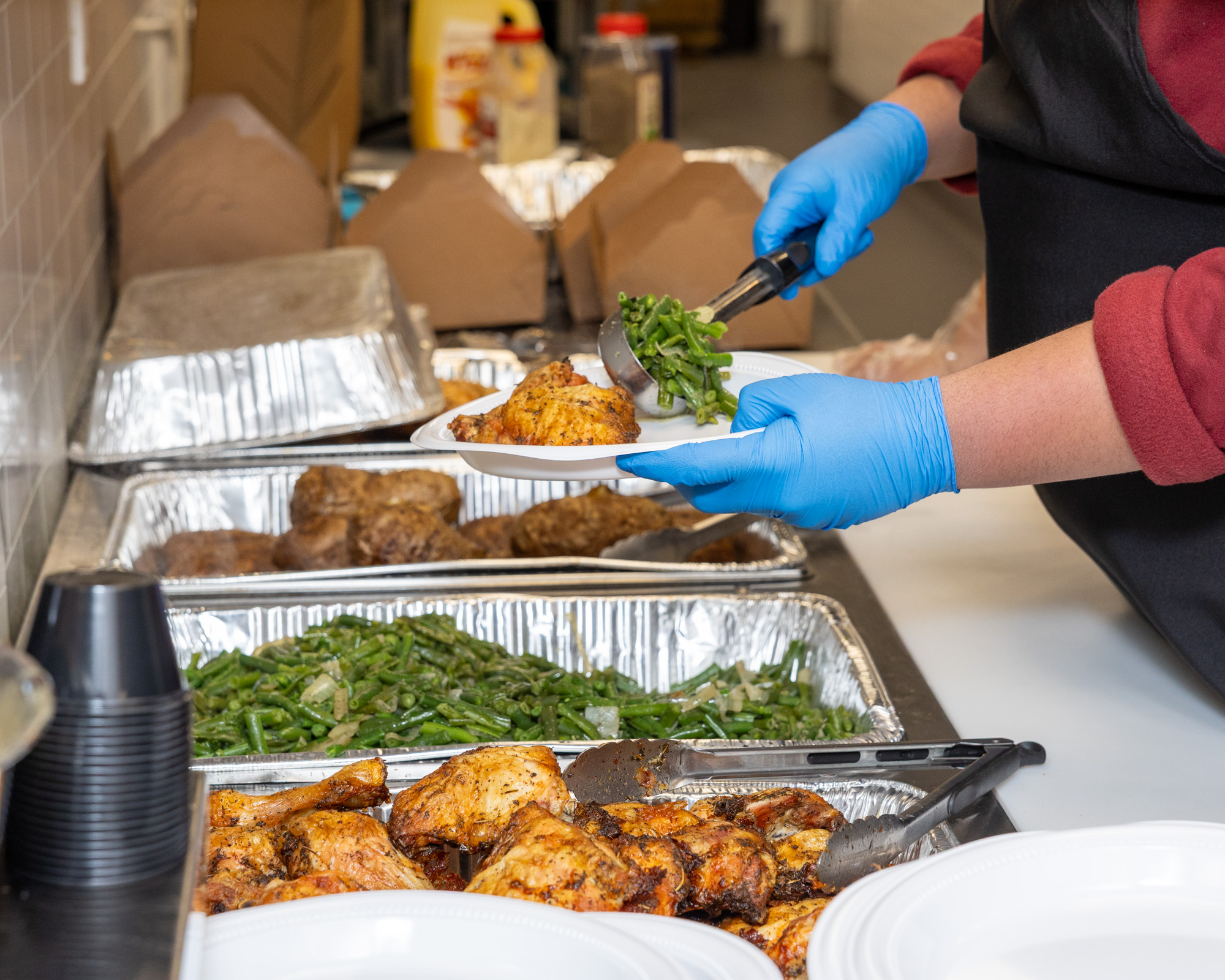 Gloved hands making a plate of chicken, beans, and potatoes in a serving kitchen.