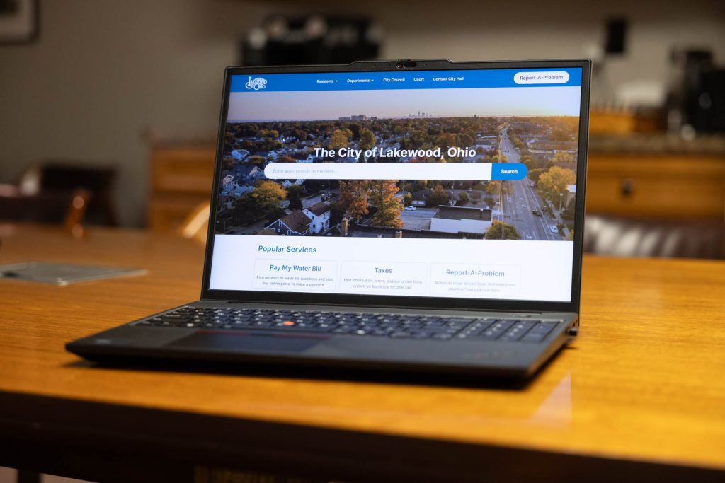 A black laptop displays the City of Lakewood website on the screen. The computer is sitting on a wood table.