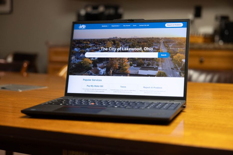 A black laptop displays the City of Lakewood website on the screen. The computer is sitting on a wood table.