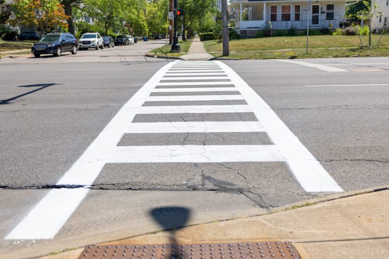A high visibility crosswalk painted on a road - two parallel white lines connected by several perpendicular painted lines.